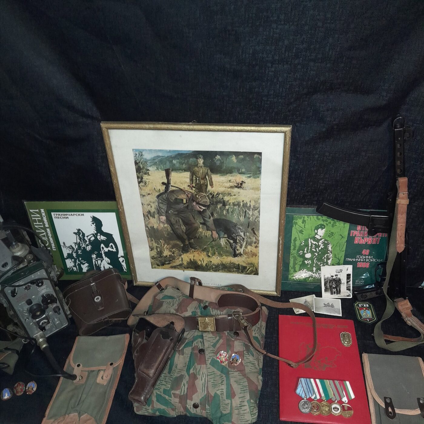 Framed period reproduction showing two border guards reading a track, with their dog, surrounded by records issued for the corps's anniversaries